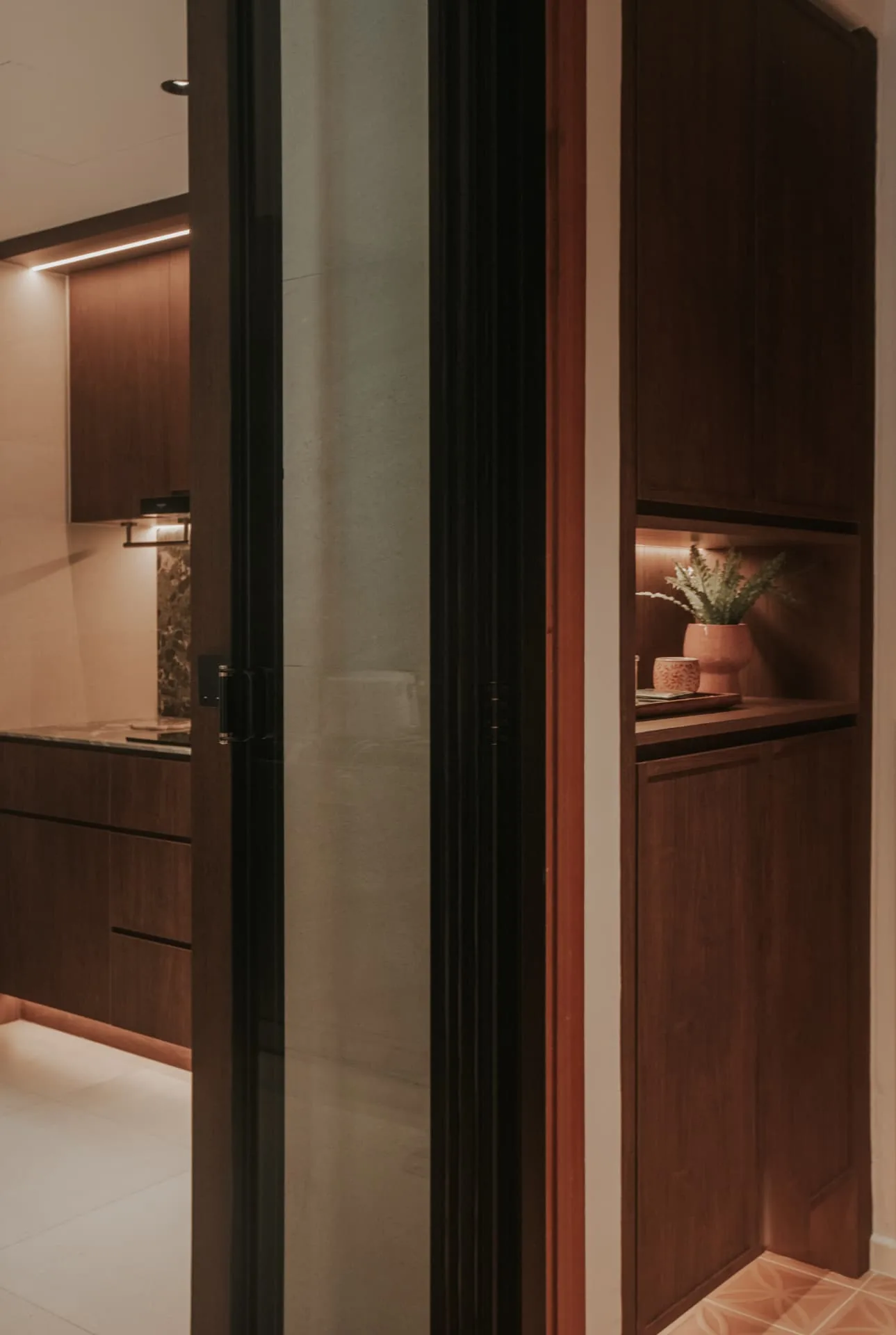Warm integrated lighting in a home interior: a lit timber display niche beside a glazed door with soft under-cabinet light in the kitchen beyond